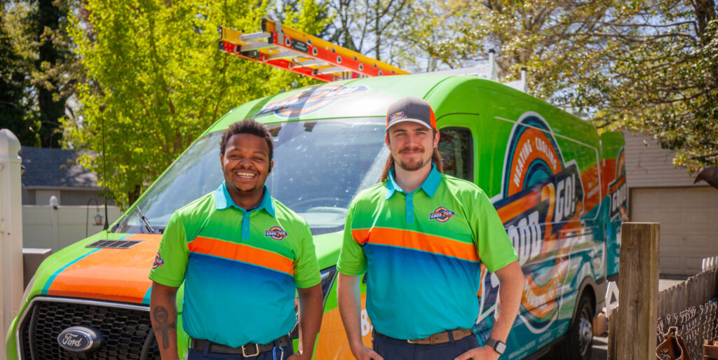 Two smiling Good2Go technicians stand in front of a brightly colored Good2Go service van, wearing matching green, blue, and orange uniforms. The branded van features bold graphics promoting heating, cooling, plumbing, and electrical services. The scene is set in a sunny residential neighborhood in New Jersey, highlighting the company’s friendly and professional team.